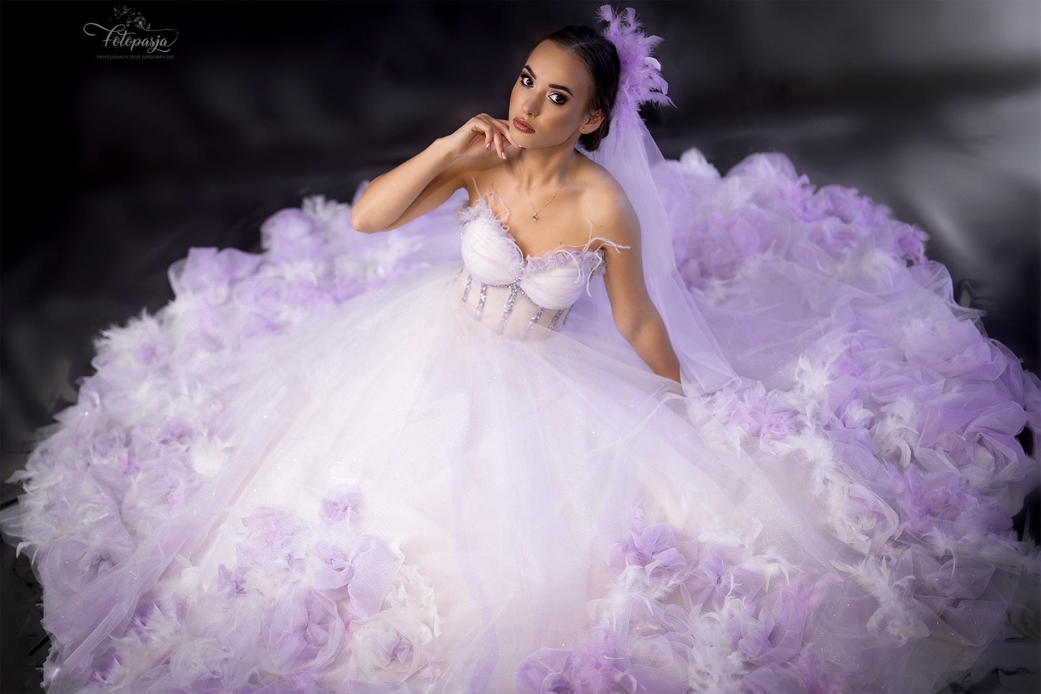 Kobieta w obszarnej, balowej sukni ślubnej w odcieniach bieli i fioletu, z dekoracjami z piór i kwiatów, pozuje na ciemnym tle.
