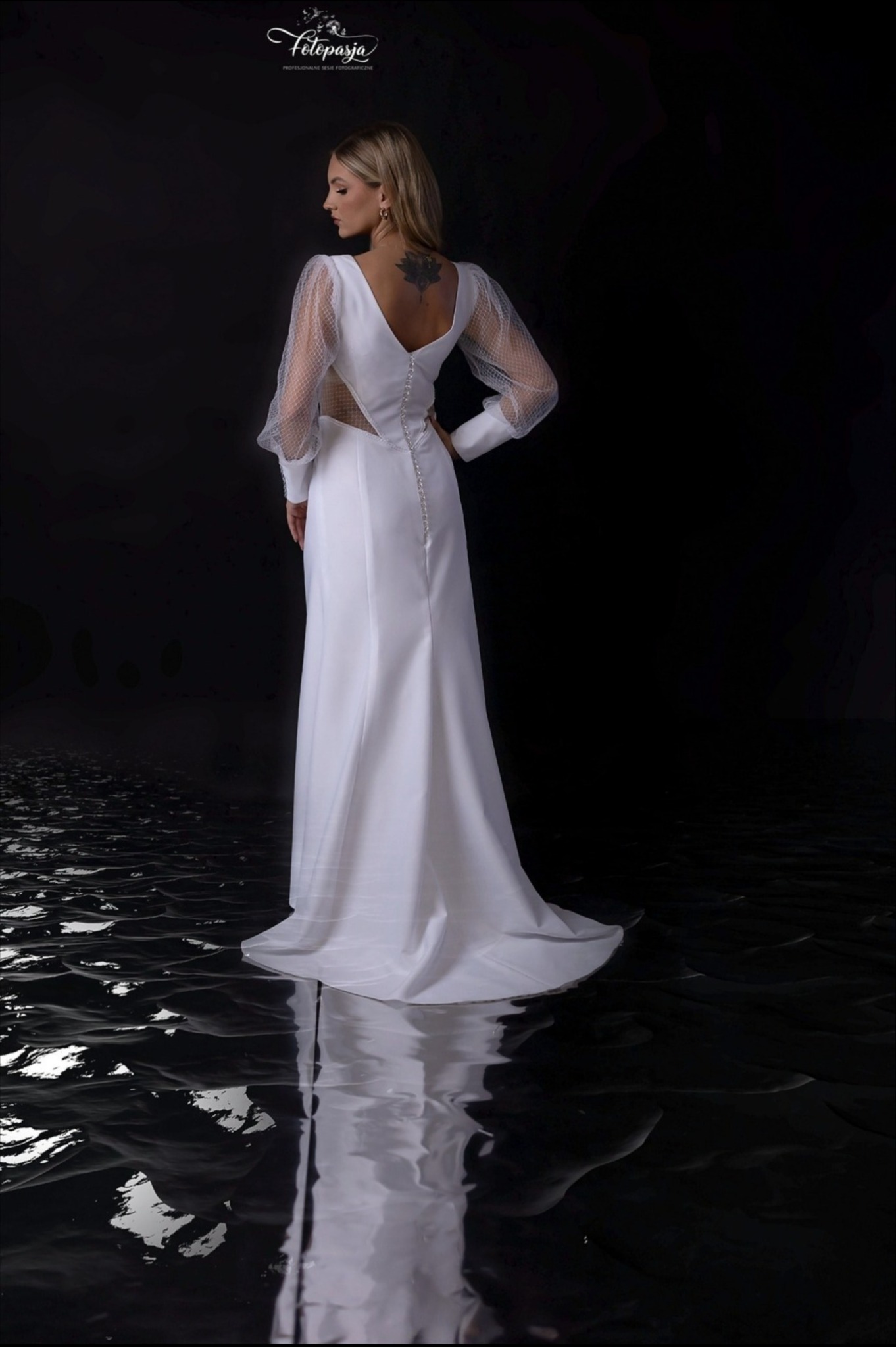 Kobieta w białej sukni z tiulowymi rękawami, widok z tyłu, z tatuażem na plecach, na ciemnym tle z odbiciem.