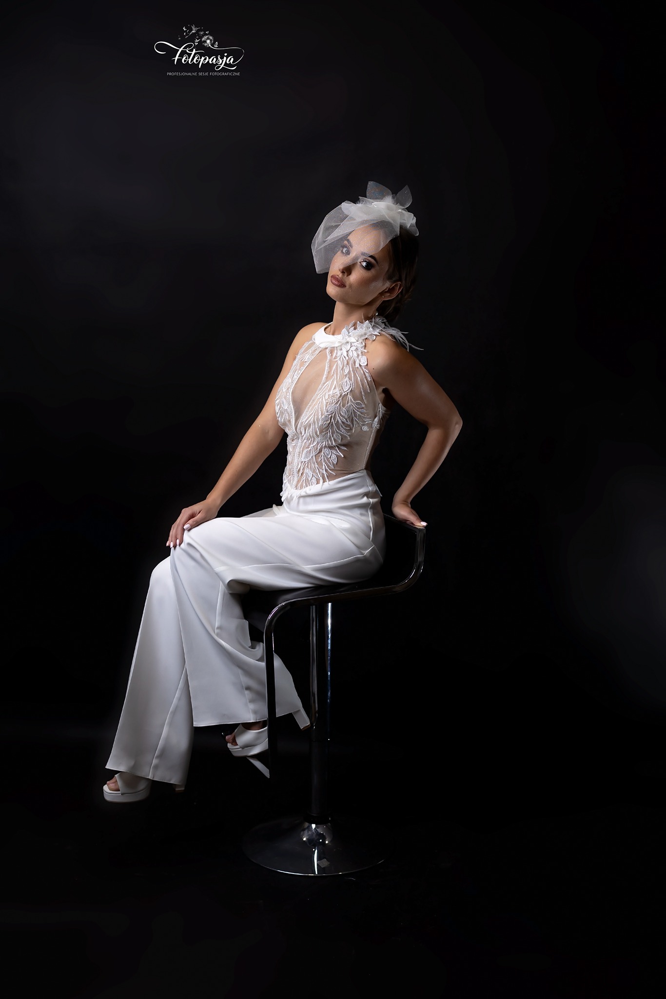 Kobieta w białym kombinezonie z koronkową górą i woalką siedzi na chromowanym stołku na czarnym tle, prezentując strój.