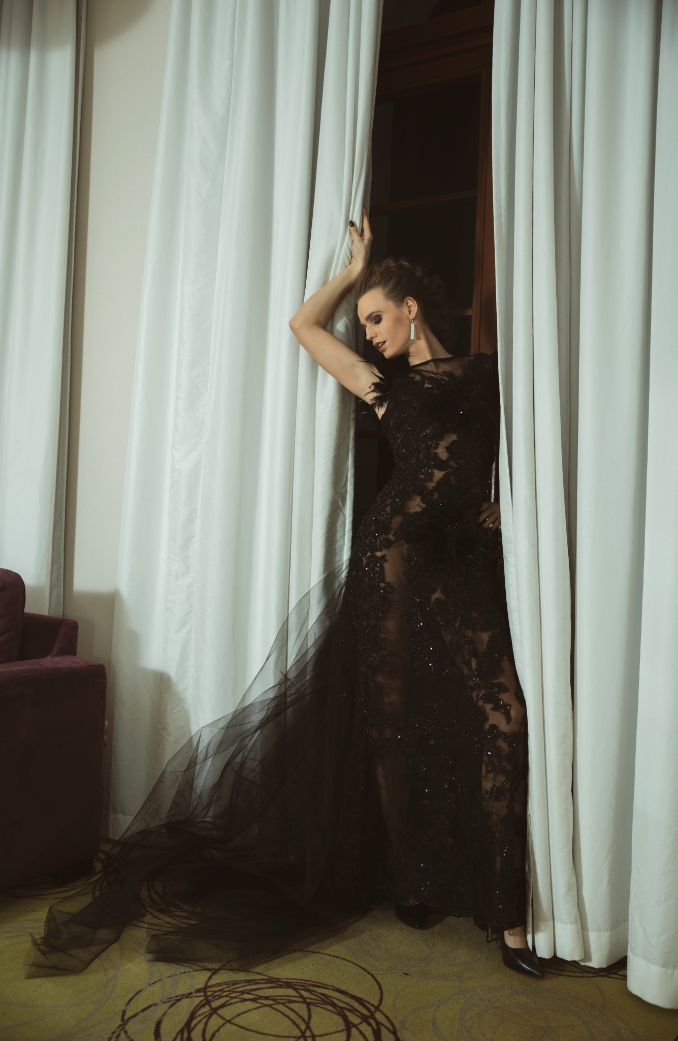 Kobieta w czarnej, koronkowej sukni wieczorowej z tiulowym trenem, pozująca przy białych zasłonach w pokoju.