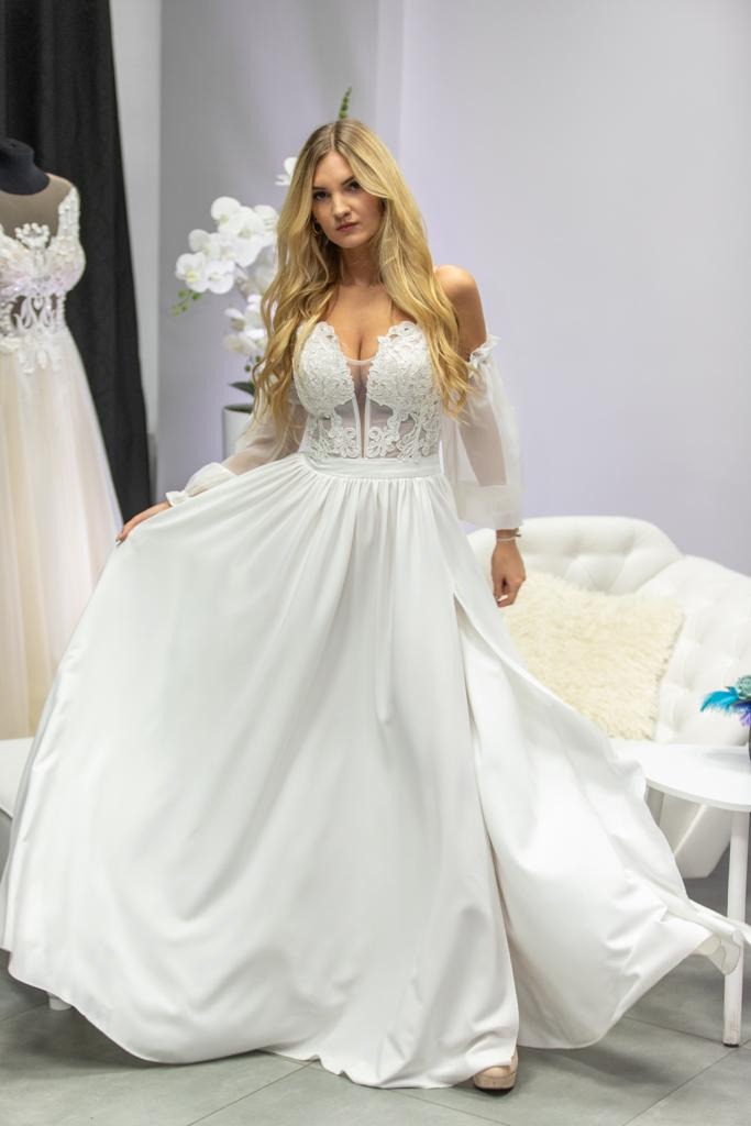 Kobieta w białej sukni ślubnej z koronkową górą i rozcięciem na nodze, pozuje w studio na tle sofy i kwiatów.