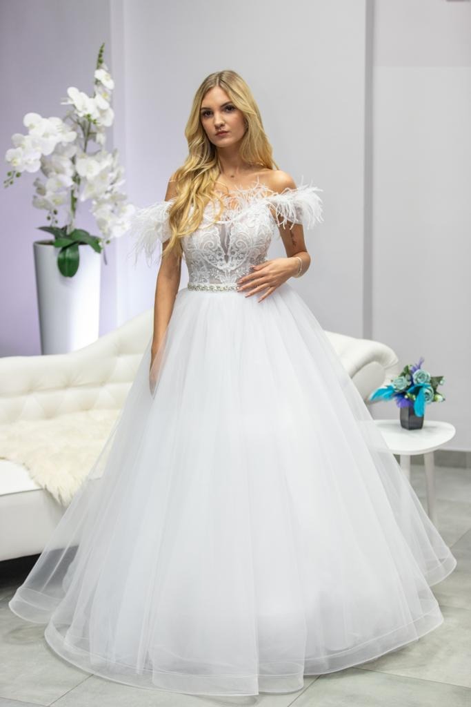 Blond modelka w białej sukni ślubnej z piórami na ramionach, koronkowym gorsetem i tiulową spódnicą, pozuje na tle białej sofy i kwiatów w salonie.