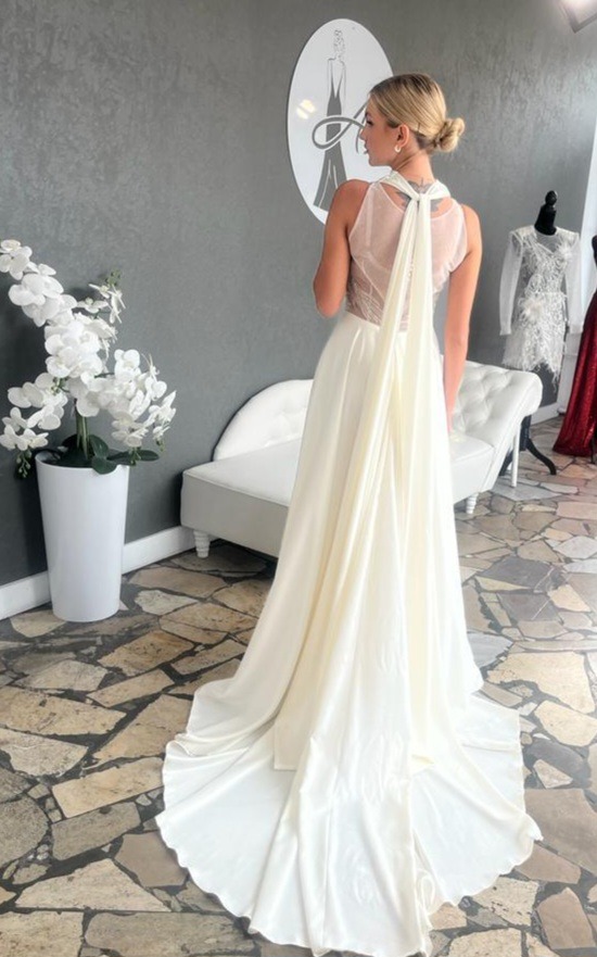 Kobieta w białej sukni ślubnej z trenem, widok z tyłu, z dekoracyjnym wiązaniem na plecach, w tle logo firmy odzieżowej i białe kwiaty w donicy.