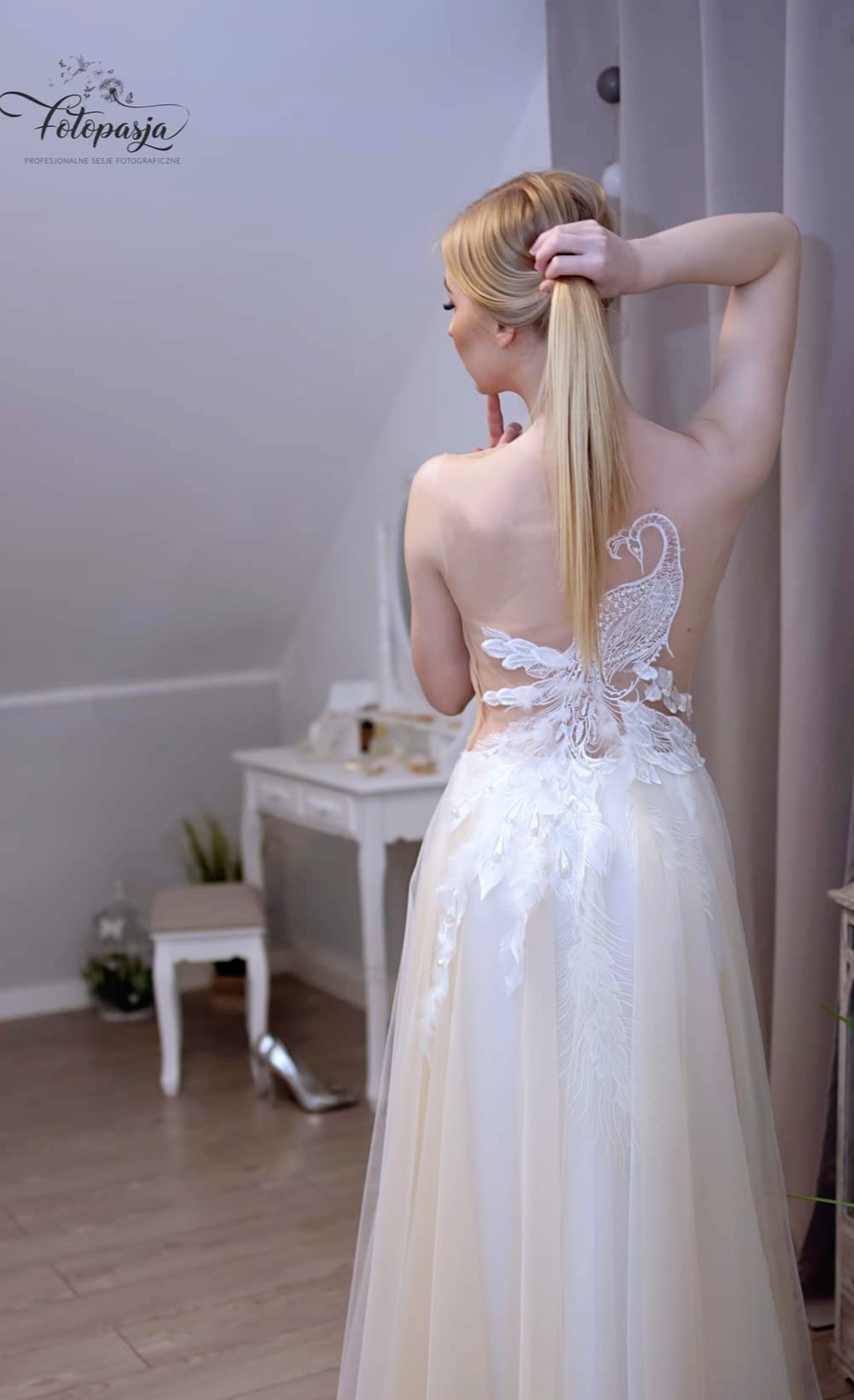 Kobieta w sukni ślubnej z odkrytymi plecami, ozdobionymi aplikacją w kształcie pawia, poprawia włosy na tle toaletki z lustrem.