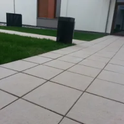 Ułożona nawierzchnia z jasnych, kwadratowych płyt betonowych prowadząca do wejścia budynku z przeszklonymi drzwiami, obok trawnik z czarnymi koszami na śmieci.