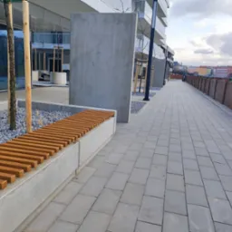 Nowoczesna przestrzeń miejska z ławką z drewnianych listew, wyłożona szarą kostką brukową, obok posadzone młode drzewa z drewnianymi podporami, w tle budynki mieszkalne i ceglany mur.