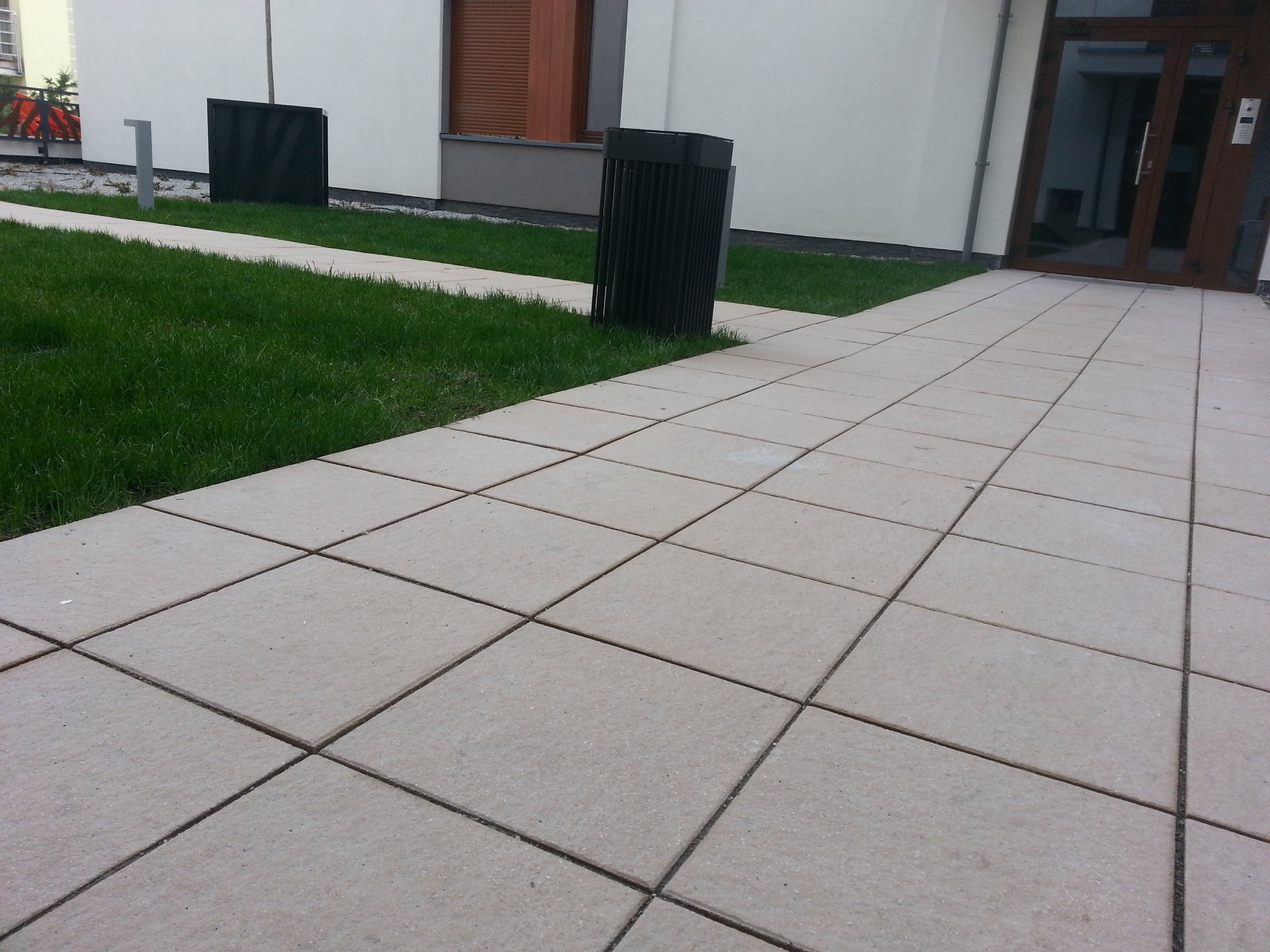 Ułożona nawierzchnia z jasnych, kwadratowych płyt betonowych prowadząca do wejścia budynku z przeszklonymi drzwiami, obok trawnik z czarnymi koszami na śmieci.