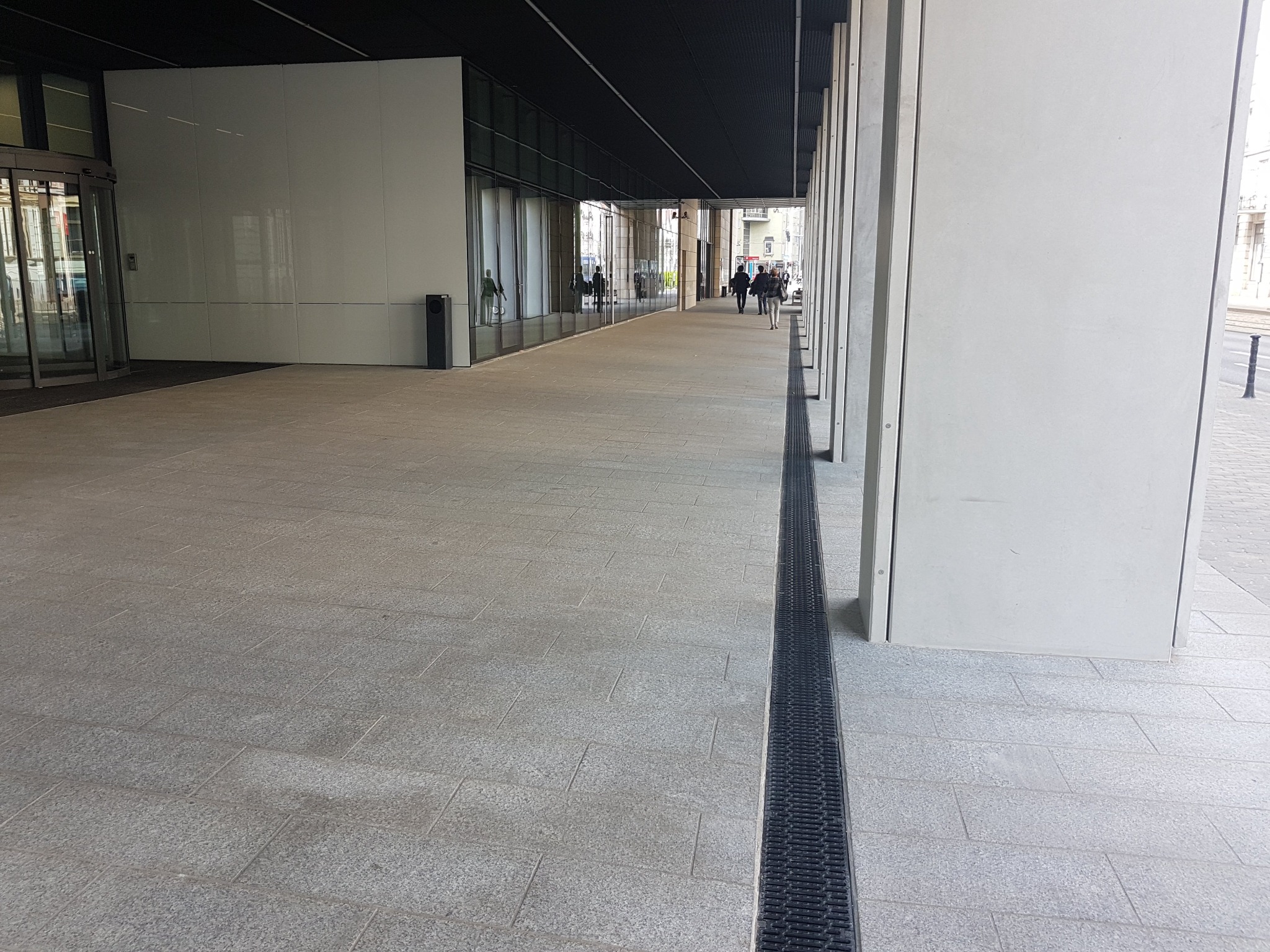 Szeroki chodnik wyłożony jasnoszarą kostką, z odpływem liniowym po prawej stronie, prowadzący do obrotowych drzwi wejściowych do budynku biurowego. W tle widoczni przechodnie.