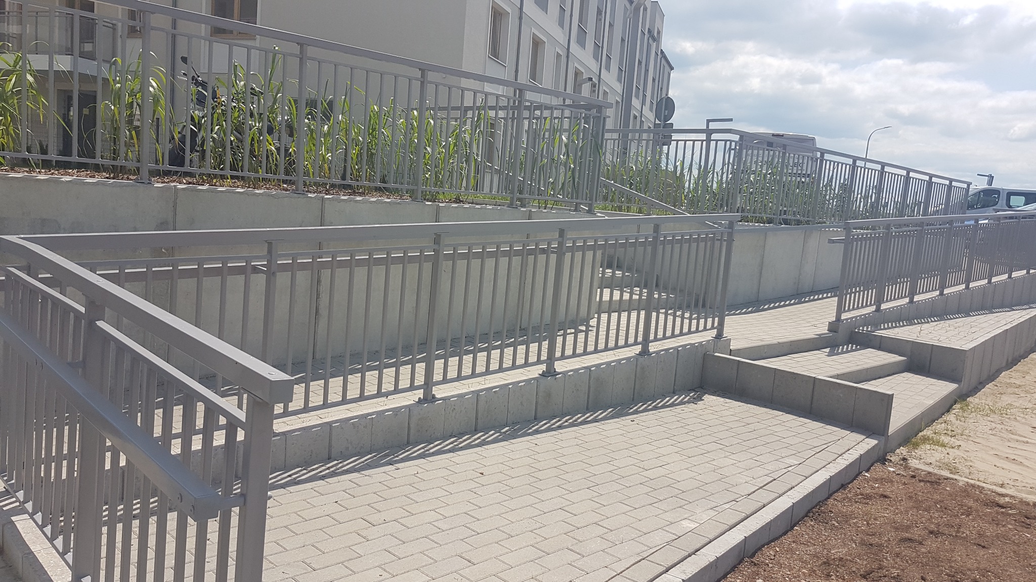 Zewnętrzne schody i podjazd dla wózków inwalidzkich, wyłożone jasną kostką brukową, z szarymi metalowymi barierkami, prowadzące do budynku z zielenią w tle.