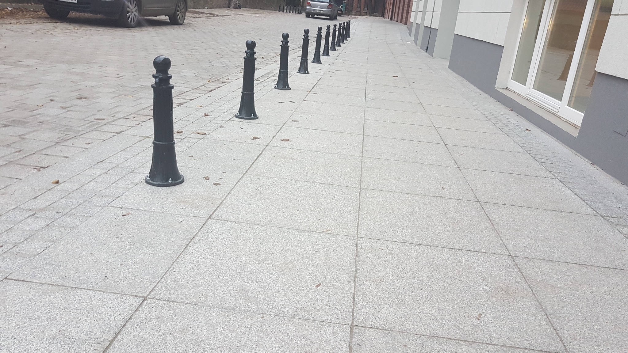 Ułożona nawierzchnia z jasnoszarej kostki brukowej z rzędem czarnych, ozdobnych słupków oddzielających chodnik od jezdni, z widocznymi samochodami w tle.
