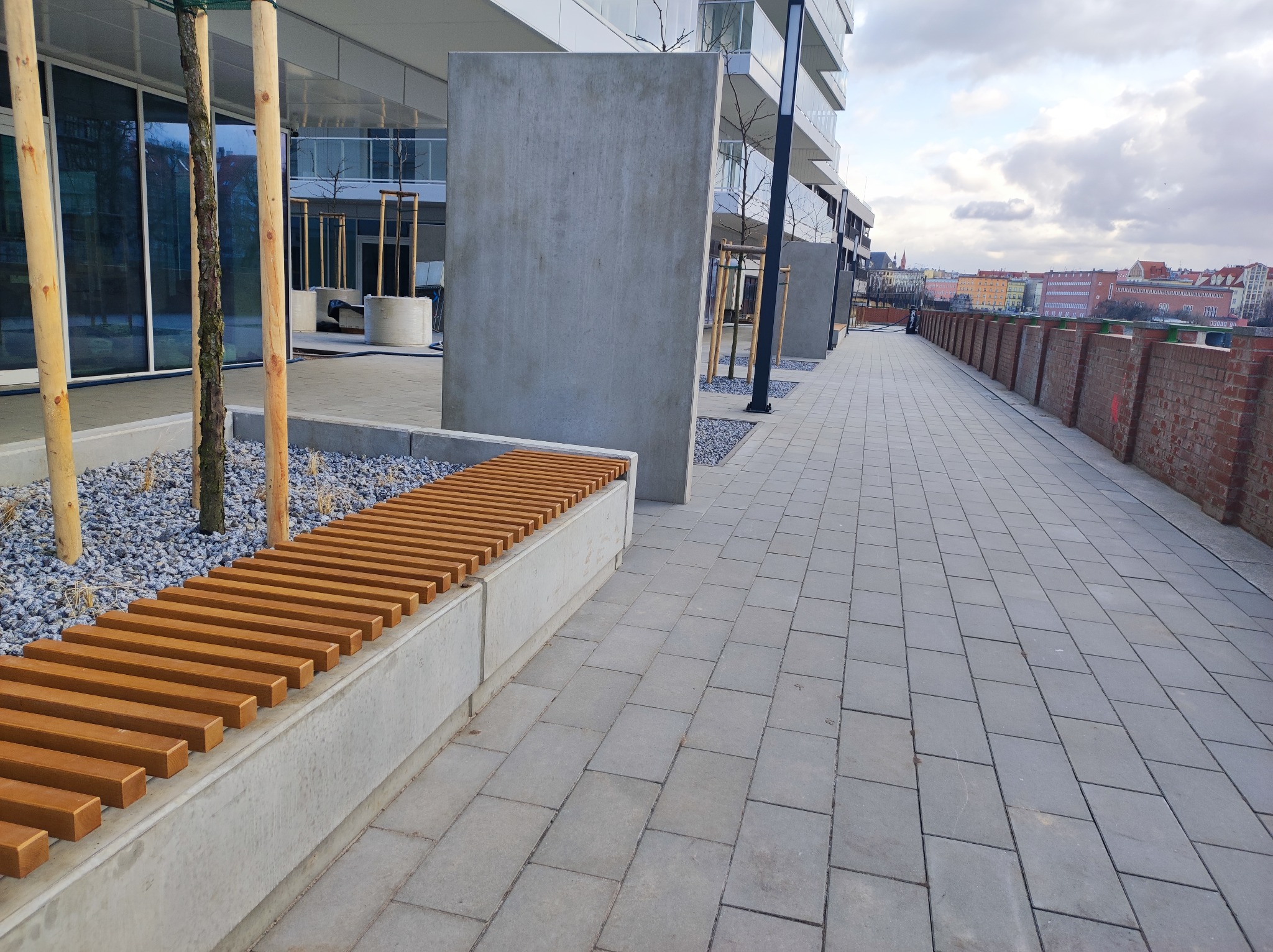 Nowoczesna przestrzeń miejska z ławką z drewnianych listew, wyłożona szarą kostką brukową, obok posadzone młode drzewa z drewnianymi podporami, w tle budynki mieszkalne i ceglany mur.