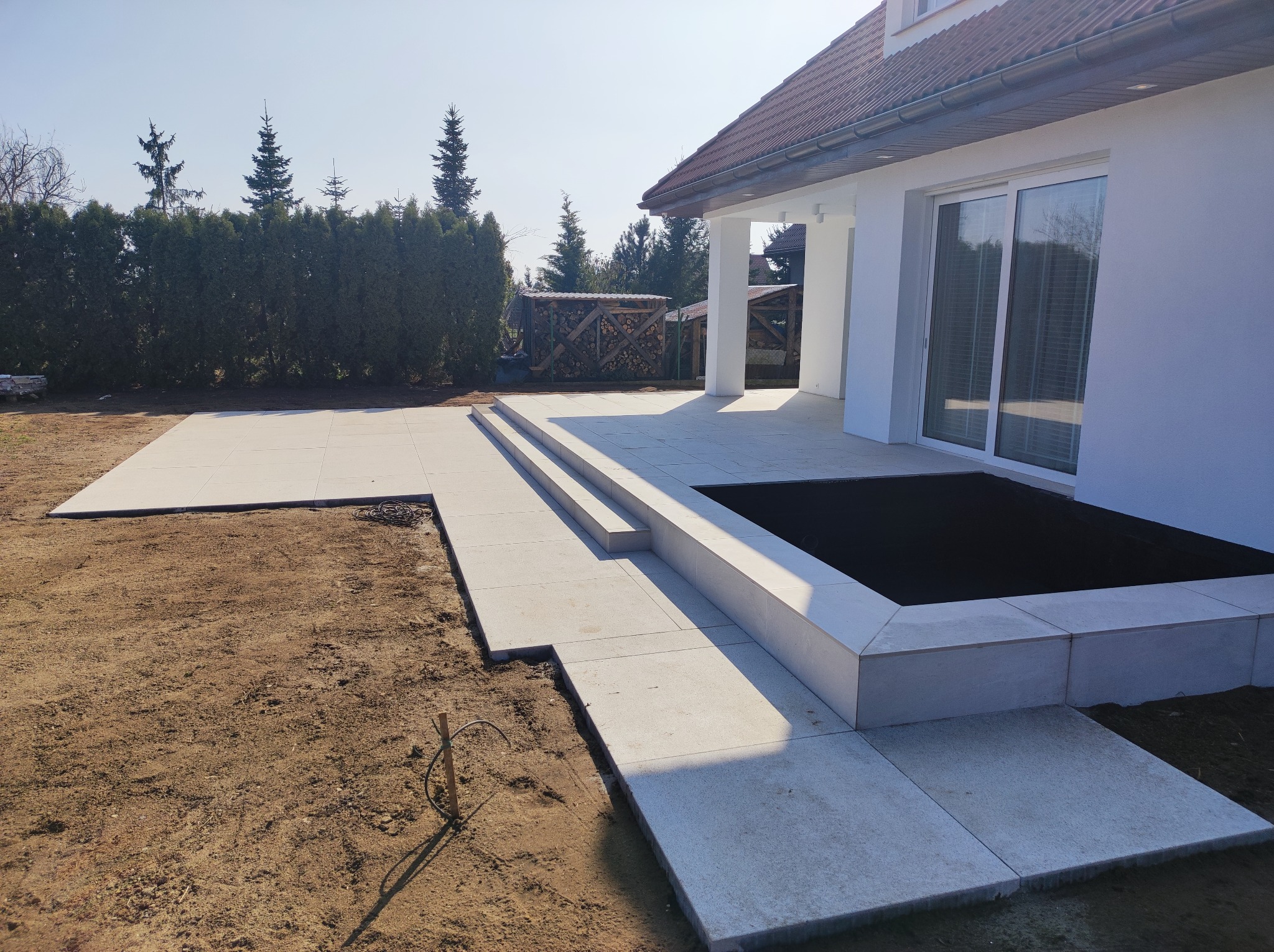 Nowoczesny taras z jasnych płyt betonowych z trzema stopniami, obok trawnik przygotowany pod zasiew, w tle biały dom z dużymi przesuwnymi drzwiami tarasowymi i zielone tuje.