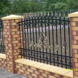 Ozdobne, kute ogrodzenie z ceglanymi słupkami i podmurówką, złote detale dekoracyjne, widok z zewnątrz.