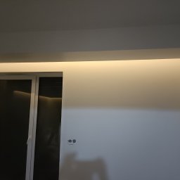 SamBud - Świeżo pomalowana ściana w jasnym odcieniu z widocznymi gniazdkami elektrycznymi i oknem z roletami, oświetlona listwą LED pod sufitem.