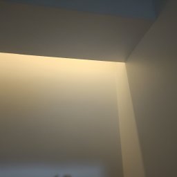 SamBud - Świeżo pomalowany narożnik sufitu i ścian w odcieniach bieli, widoczne delikatne przejścia kolorystyczne pod wpływem oświetlenia.