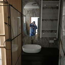 SamBud - Nowoczesna łazienka z kabiną prysznicową typu walk-in, owalnym lustrem i grzejnikiem drabinkowym na tle ściany imitującej marmur, z odbiciem fotografa w lustrze.