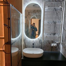 SamBud - Nowoczesna łazienka z marmurową ścianą, owalnym lustrem z podświetleniem LED, białą umywalką nablatową na czarnej szafce i grzejnikiem drabinkowym.