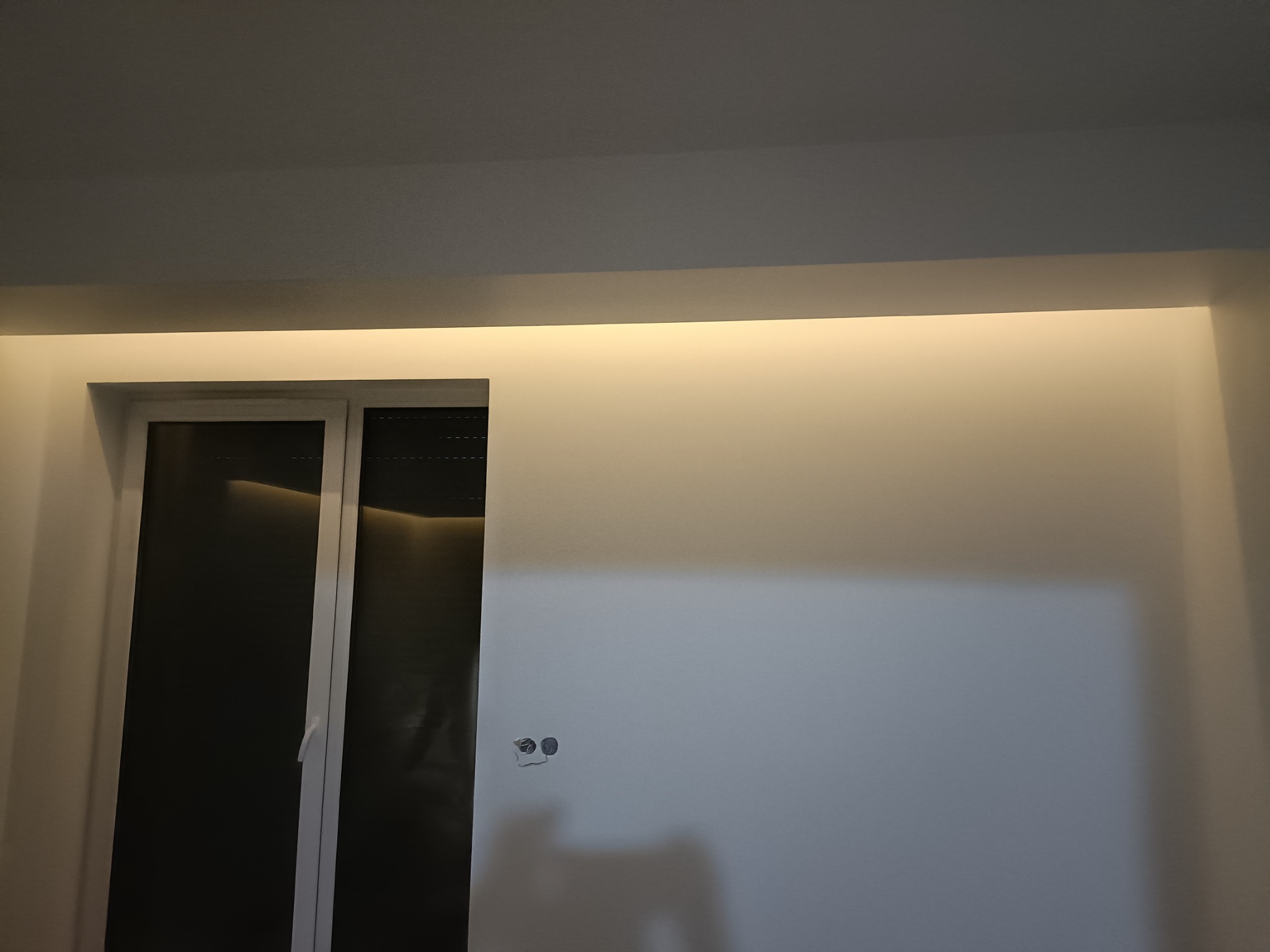 Świeżo pomalowana ściana w jasnym odcieniu z widocznymi gniazdkami elektrycznymi i oknem z roletami, oświetlona listwą LED pod sufitem.