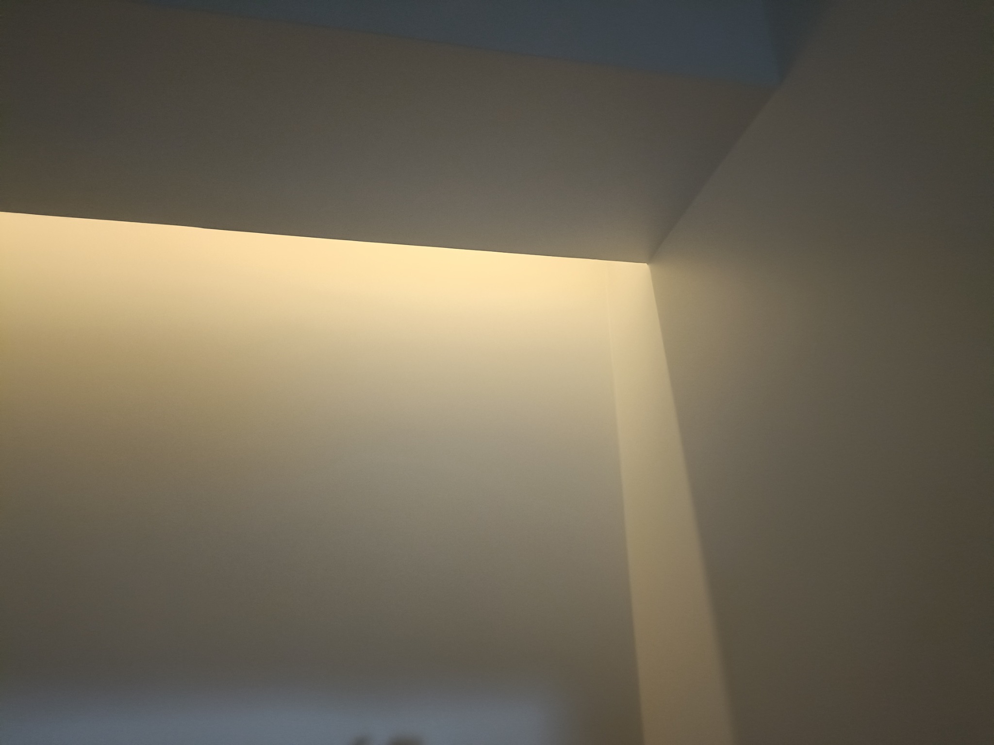 Świeżo pomalowany narożnik sufitu i ścian w odcieniach bieli, widoczne delikatne przejścia kolorystyczne pod wpływem oświetlenia.