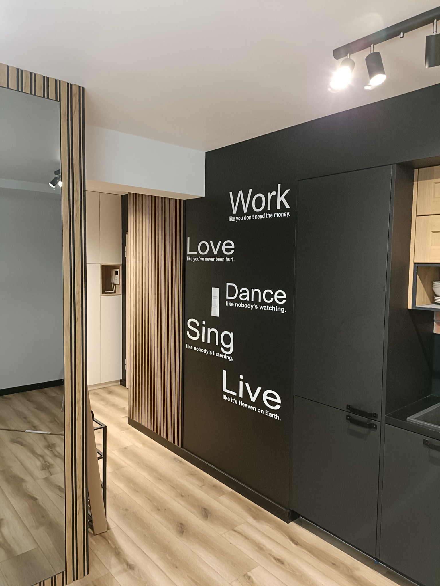 Nowoczesne wnętrze z drewnianymi lamelami na ścianie, czarną zabudową kuchenną z białymi napisami motywacyjnymi i oświetleniem punktowym, całość utrzymana w jasnej kolorystyce podłogi.
