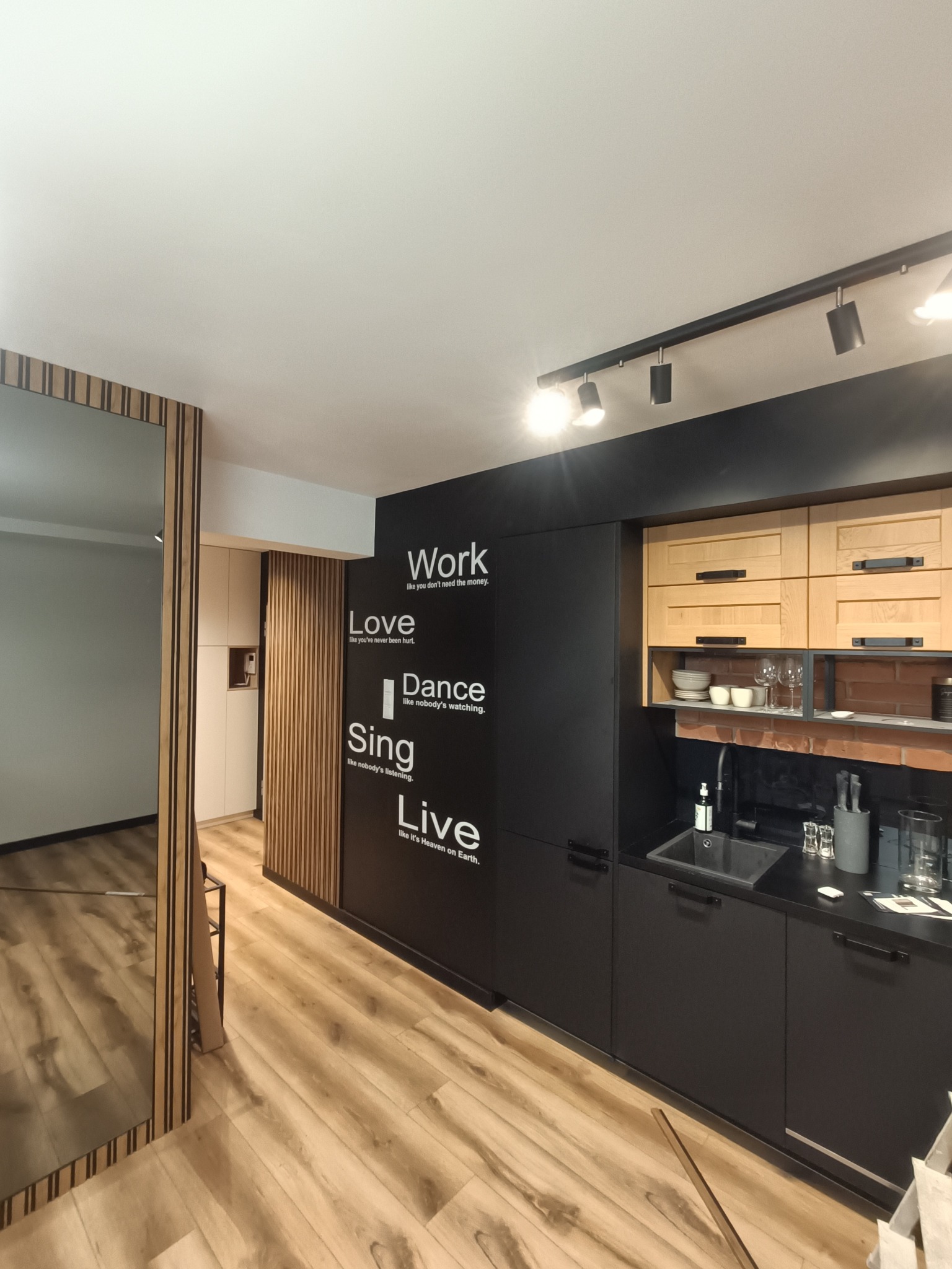 Nowoczesna kuchnia z czarnymi szafkami, drewnianymi akcentami i ścianą z napisem motywacyjnym, oświetlona punktowymi lampami na szynie.