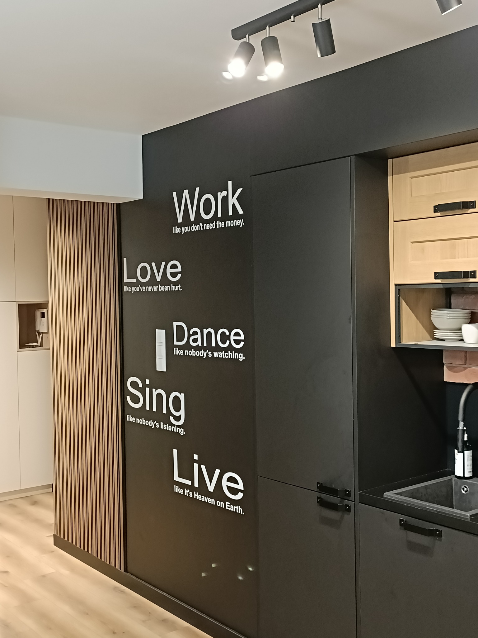 Nowoczesna kuchnia z czarnymi szafkami ozdobionymi białymi napisami motywacyjnymi, drewnianymi akcentami i oświetleniem punktowym.