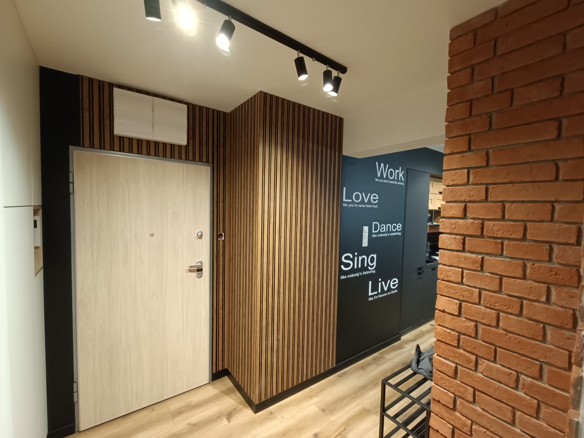 Nowoczesne wnętrze mieszkania z drewnianą okładziną ścienną, jasnymi panelami podłogowymi, ceglanym narożnikiem i drzwiami wejściowymi z jasnego drewna, oświetlone reflektorami sufitowymi.