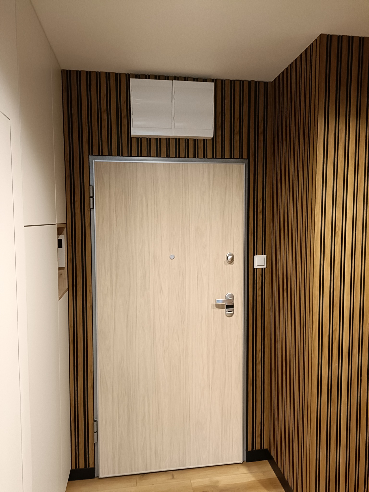 Jasne, drewnopodobne drzwi wejściowe z srebrną klamką i wizjerem, osadzone w ścianie z dekoracyjnych, pionowych paneli drewnianych, obok biała szafa wnękowa.