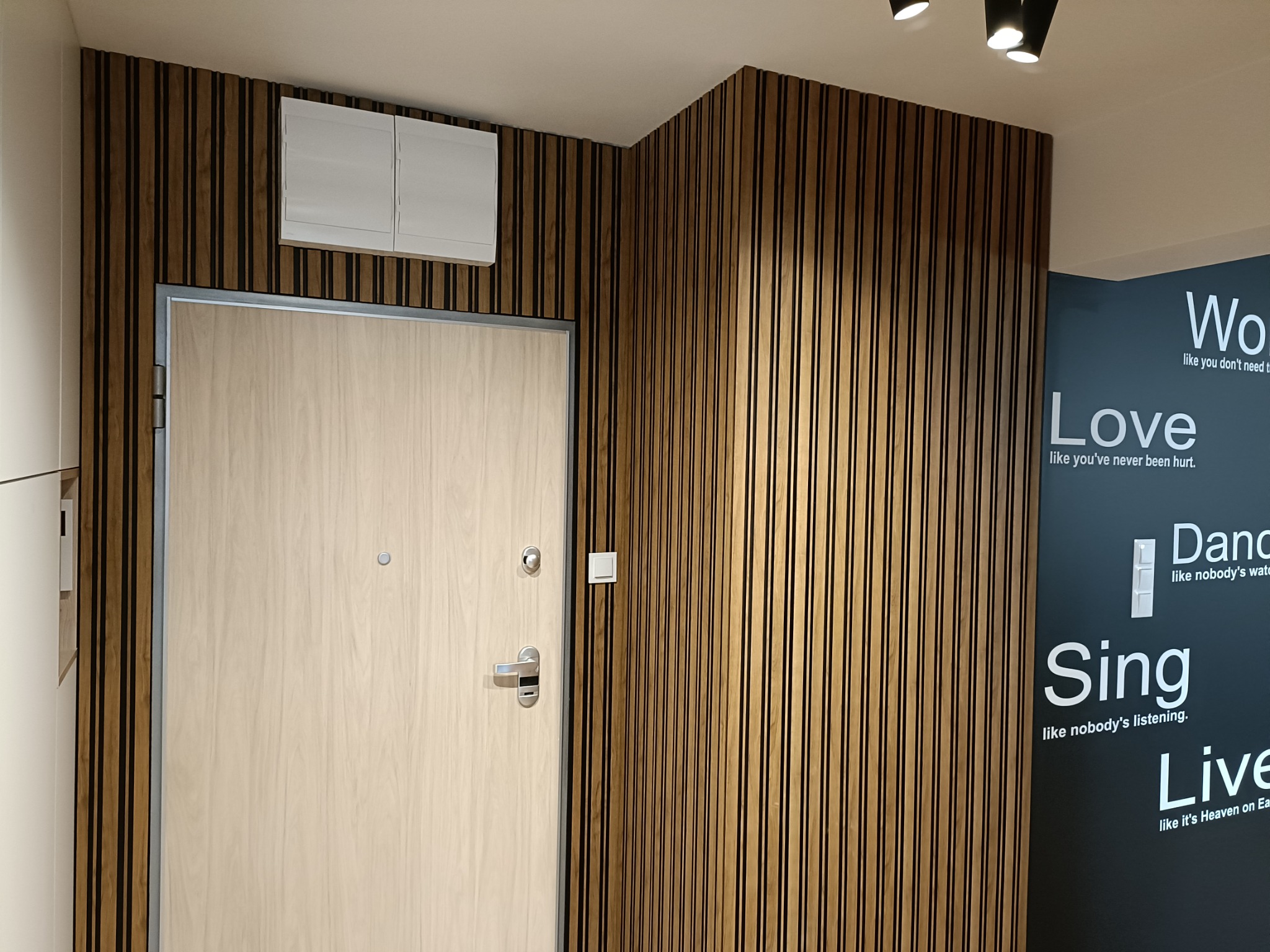 Jasne drewniane drzwi wejściowe z srebrną klamką i wizjerem, zamontowane w ścianie z pionowych drewnianych paneli, nad drzwiami biała szafka, na ścianie obok dekoracyjne napisy w języku angielskim.