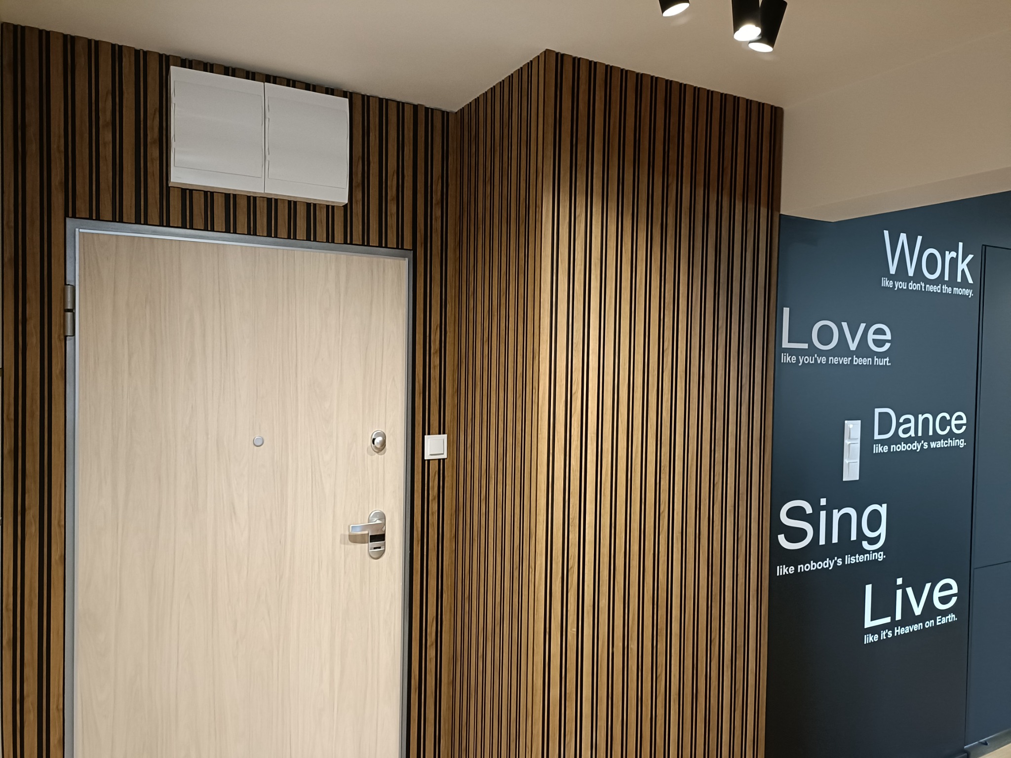 Jasne drewniane drzwi wejściowe z metalową ościeżnicą, zamontowane w ścianie z dekoracyjnych paneli z ciemnego drewna, obok ściana z napisem 'Work, Love, Dance, Sing, Live'.