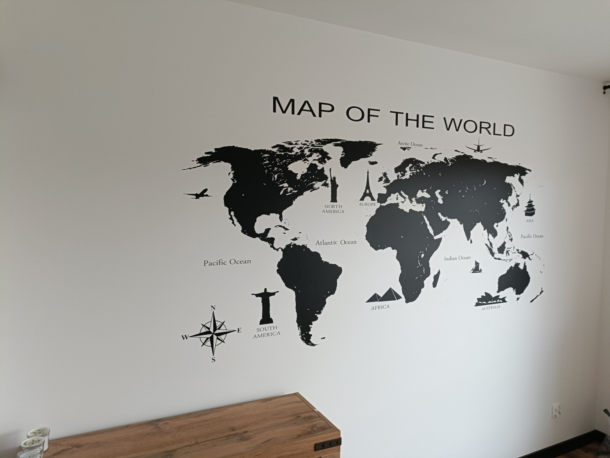 Ściana z naklejoną czarną mapą świata z nazwami kontynentów i oceanów, poniżej widoczna drewniana skrzynia.