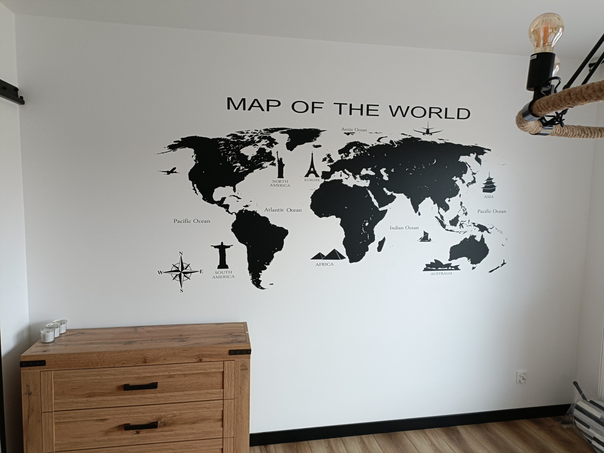 Ściana pokoju z naklejoną czarną mapą świata z zaznaczonymi kontynentami i charakterystycznymi budowlami, drewniana komoda z trzema szufladami w rogu, lampa wisząca z żarówką Edisona