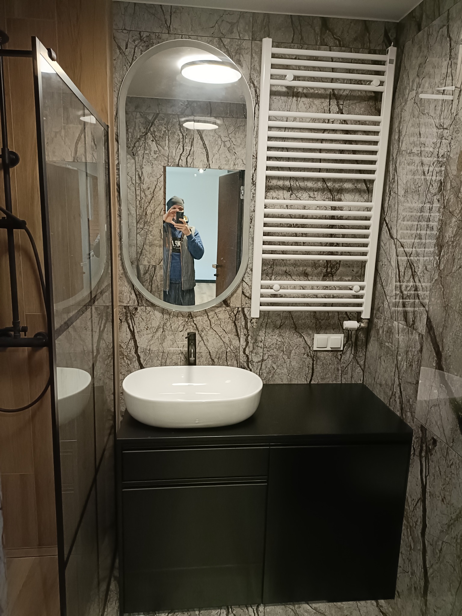 Nowoczesna łazienka z umywalką nablatową, czarną szafką pod umywalką, grzejnikiem drabinkowym i lustrem owalnym, wykończona płytkami imitującymi marmur.