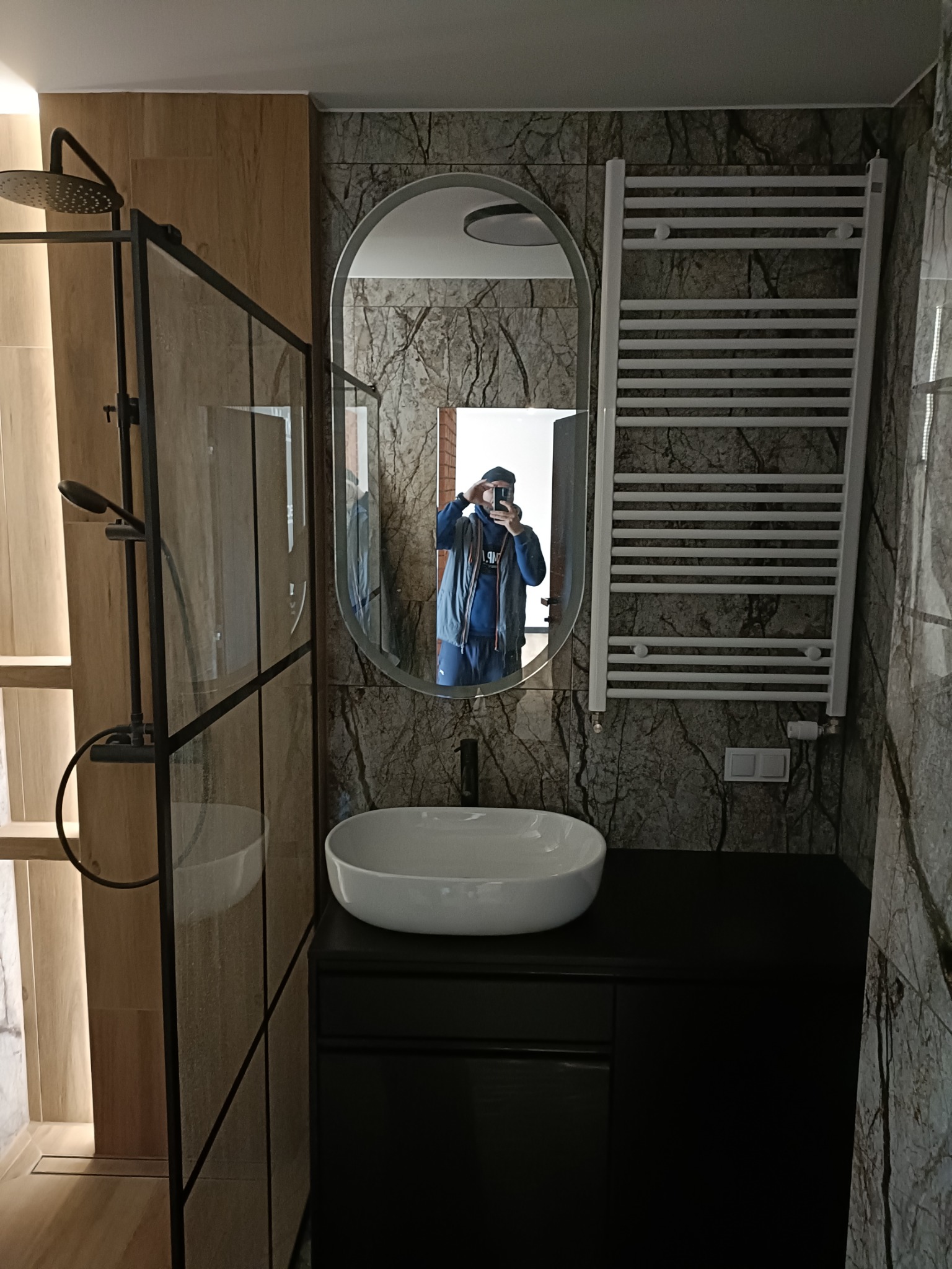 Nowoczesna łazienka z kabiną prysznicową typu walk-in, owalnym lustrem i grzejnikiem drabinkowym na tle ściany imitującej marmur, z odbiciem fotografa w lustrze.