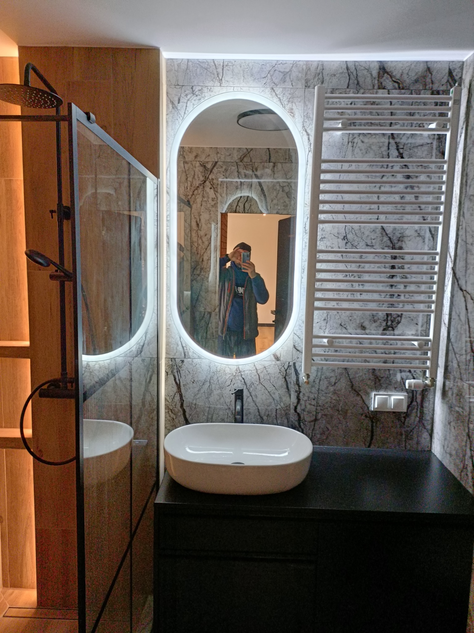 Nowoczesna łazienka z marmurową ścianą, owalnym lustrem z podświetleniem LED, białą umywalką nablatową na czarnej szafce i grzejnikiem drabinkowym.