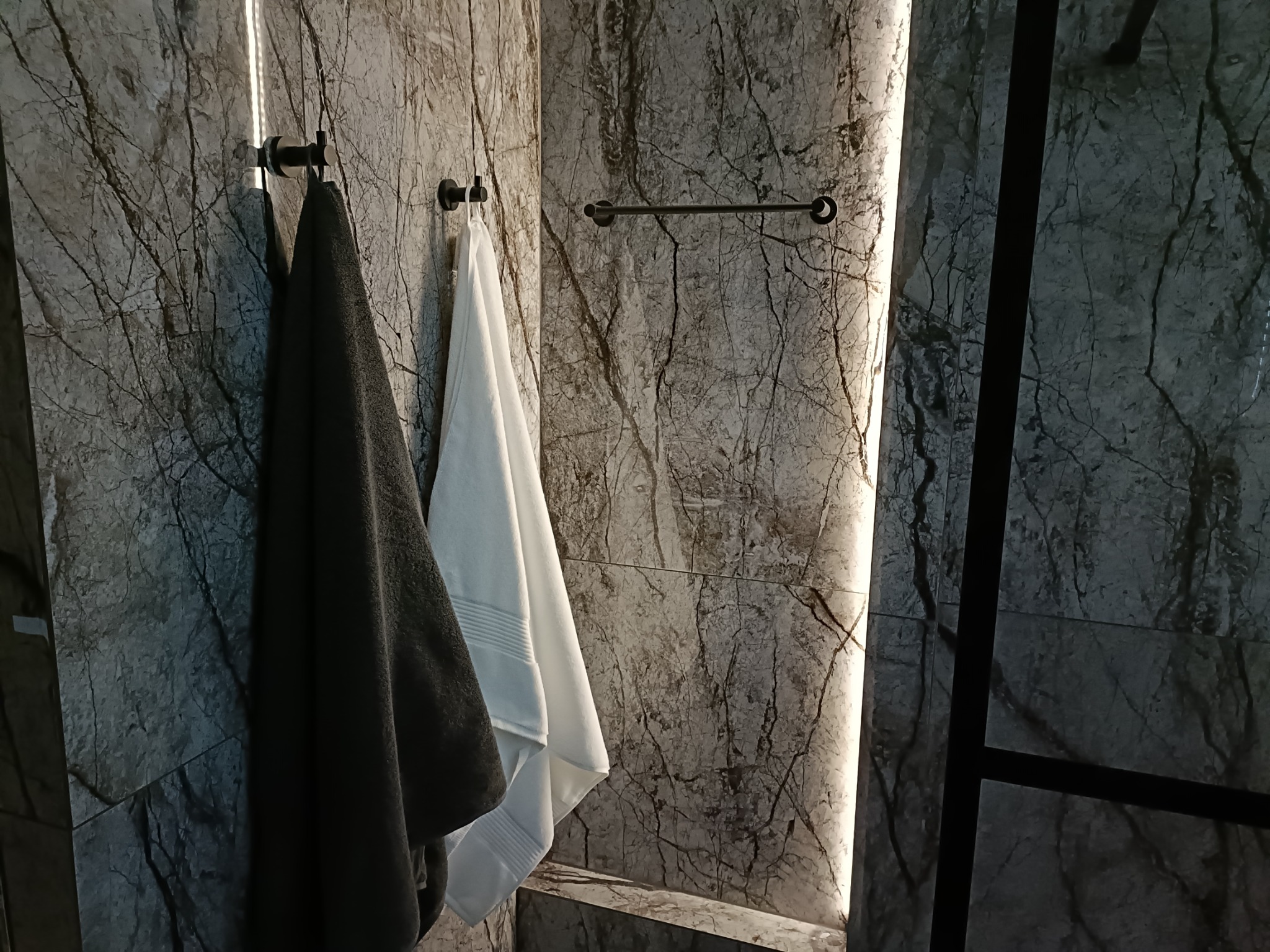 Nowoczesna łazienka wykończona płytami imitującymi marmur, z czarnymi haczykami i wieszakiem na ręczniki, na których wiszą szary i biały ręcznik.