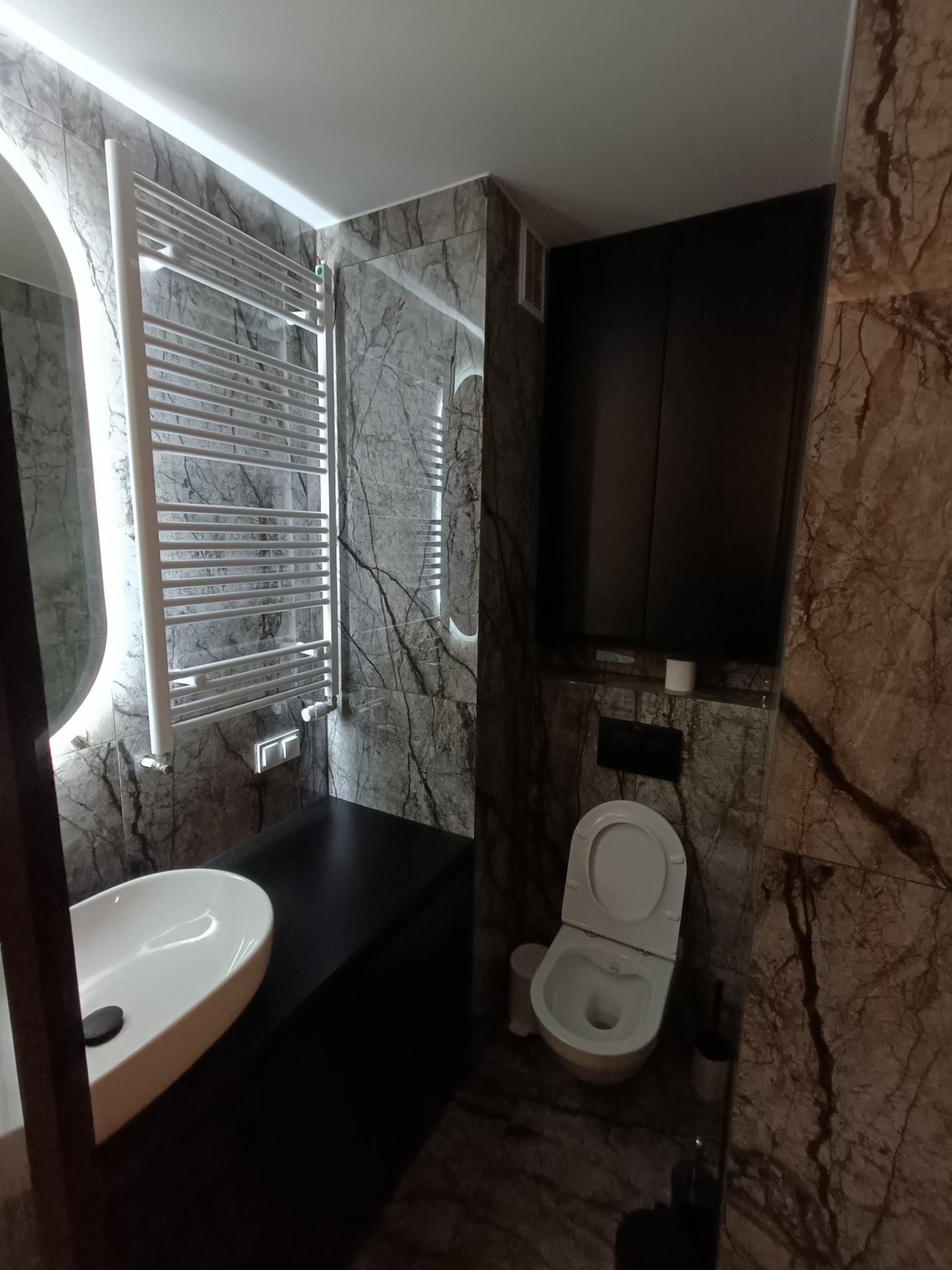 Nowoczesna łazienka z marmurowymi płytkami, podświetlanym lustrem, białą umywalką nablatową i grzejnikiem drabinkowym.