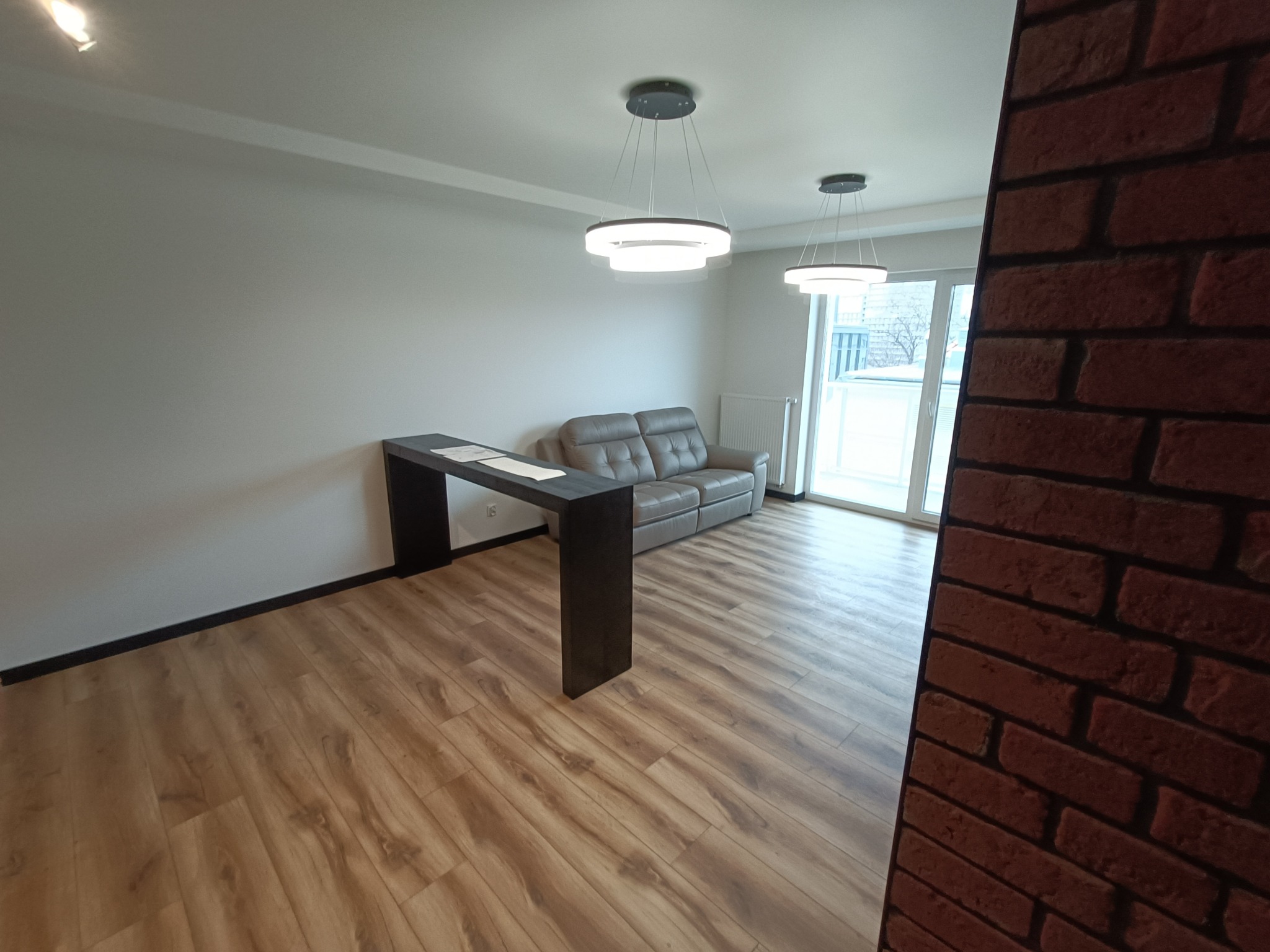 Jasne wnętrze po remoncie: drewniana podłoga, szara sofa, nowoczesne lampy wiszące i ściana z imitacji czerwonej cegły.