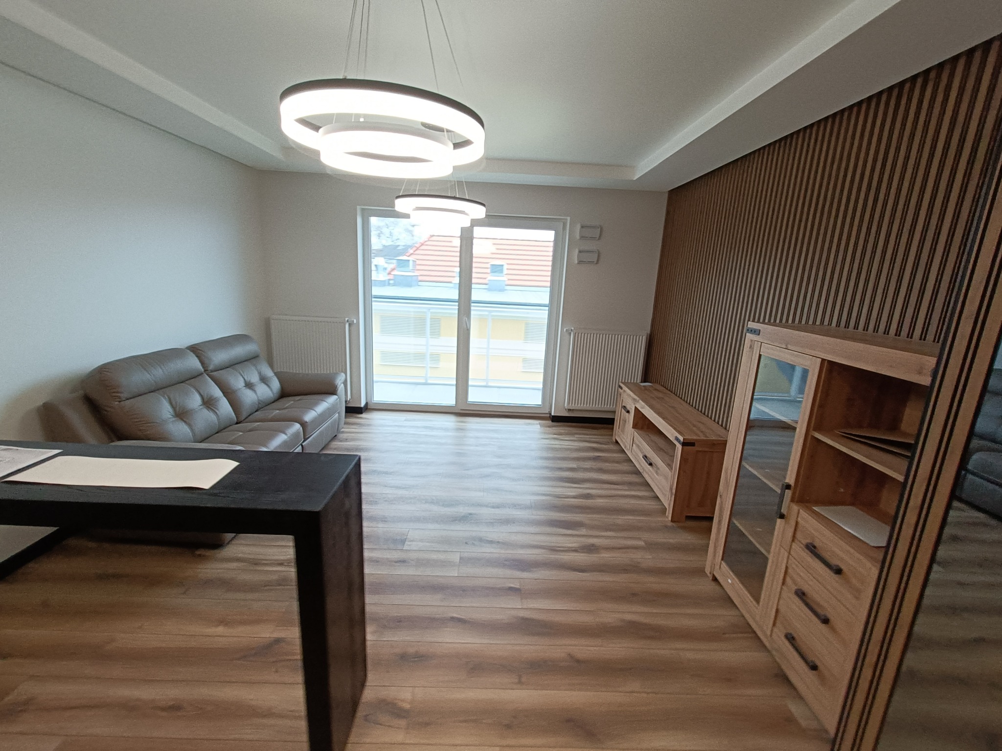 Jasne wnętrze salonu z drewnianą podłogą, szarą sofą, drewnianą ścianą z lamelami i nowoczesnym oświetleniem sufitowym.