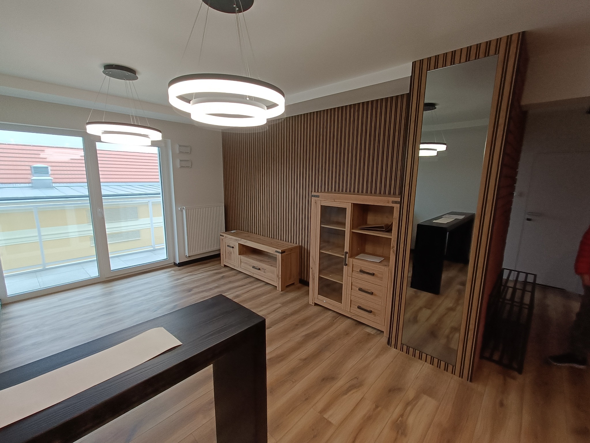 Jasne wnętrze salonu z drewnianą podłogą, meblami z jasnego drewna, lamelami na ścianie i nowoczesnym oświetleniem sufitowym. Duże okno balkonowe wpuszcza naturalne światło.