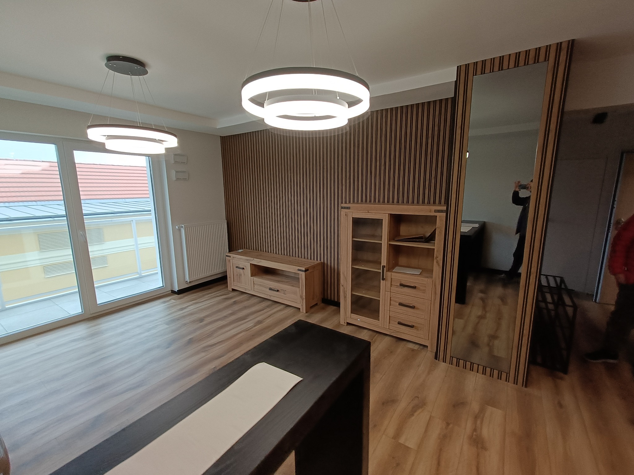 Jasne wnętrze salonu po remoncie, z drewnianą boazerią pionową, drewnianymi meblami, dużym lustrem w drewnianej ramie i nowoczesnymi lampami sufitowymi.