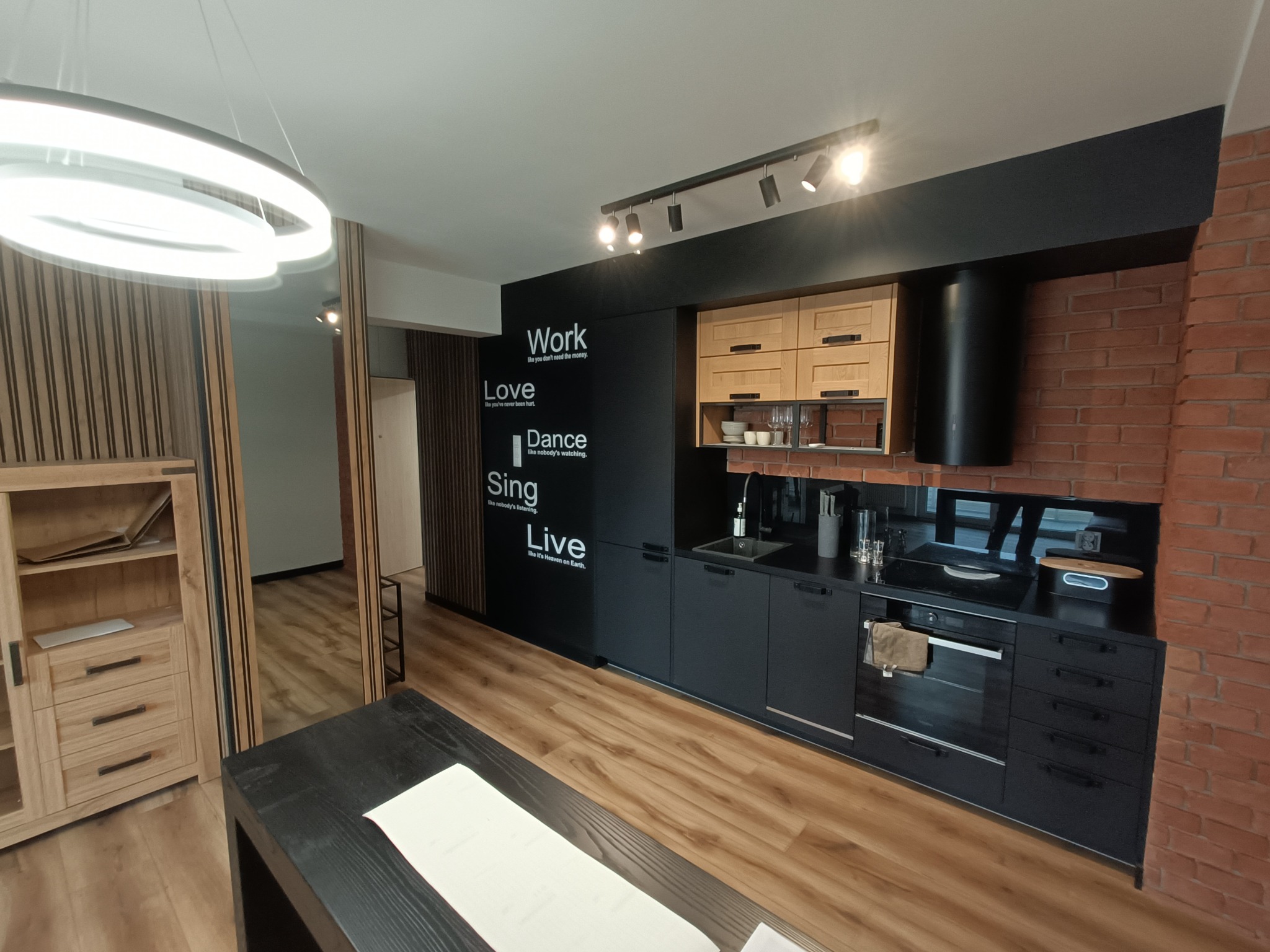Nowoczesna kuchnia w stylu loft z czarnymi szafkami, ceglaną ścianą i drewnianymi akcentami, oświetlona lampami punktowymi i okrągłą lampą wiszącą, z napisem motywacyjnym na ścianie.