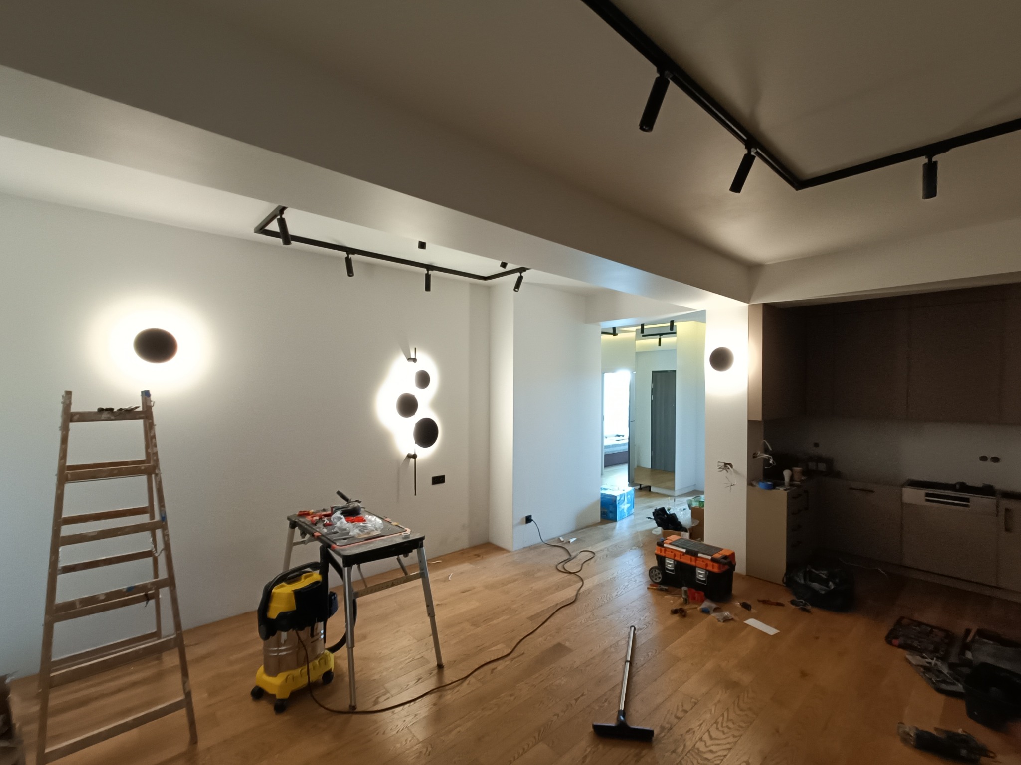 Wnętrze w trakcie prac: drewniana podłoga, drabina oparta o ścianę z okrągłymi lampami, narzędzia na stole warsztatowym, widoczne kable i lampy sufitowe na szynoprzewodach.