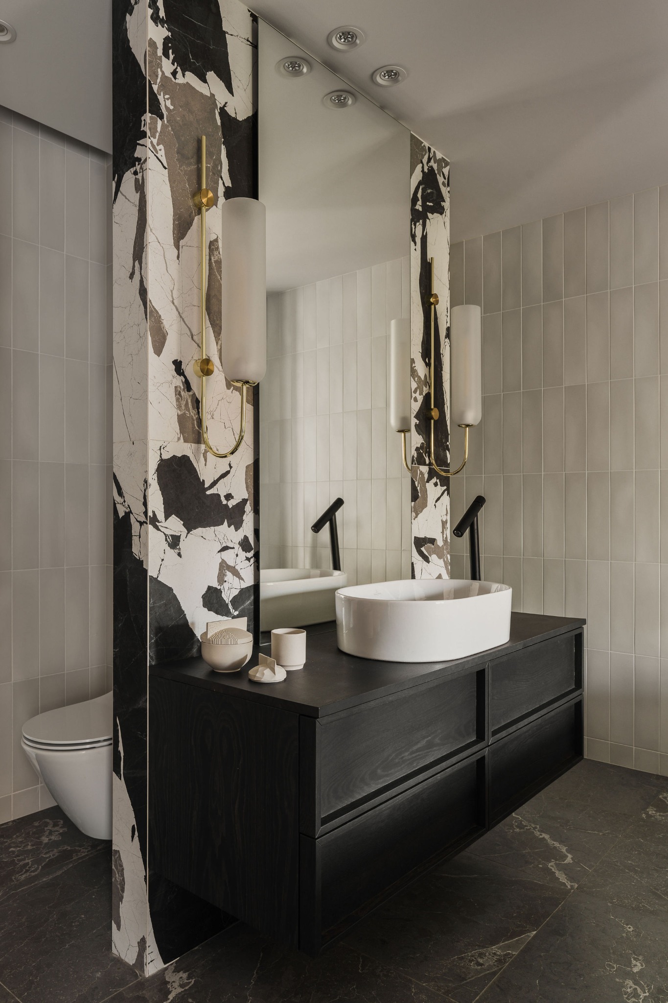 Nowoczesna łazienka z czarną szafką pod umywalką, białą owalną umywalką nablatową, złotymi kinkietami i marmurowym akcentem na ścianie.