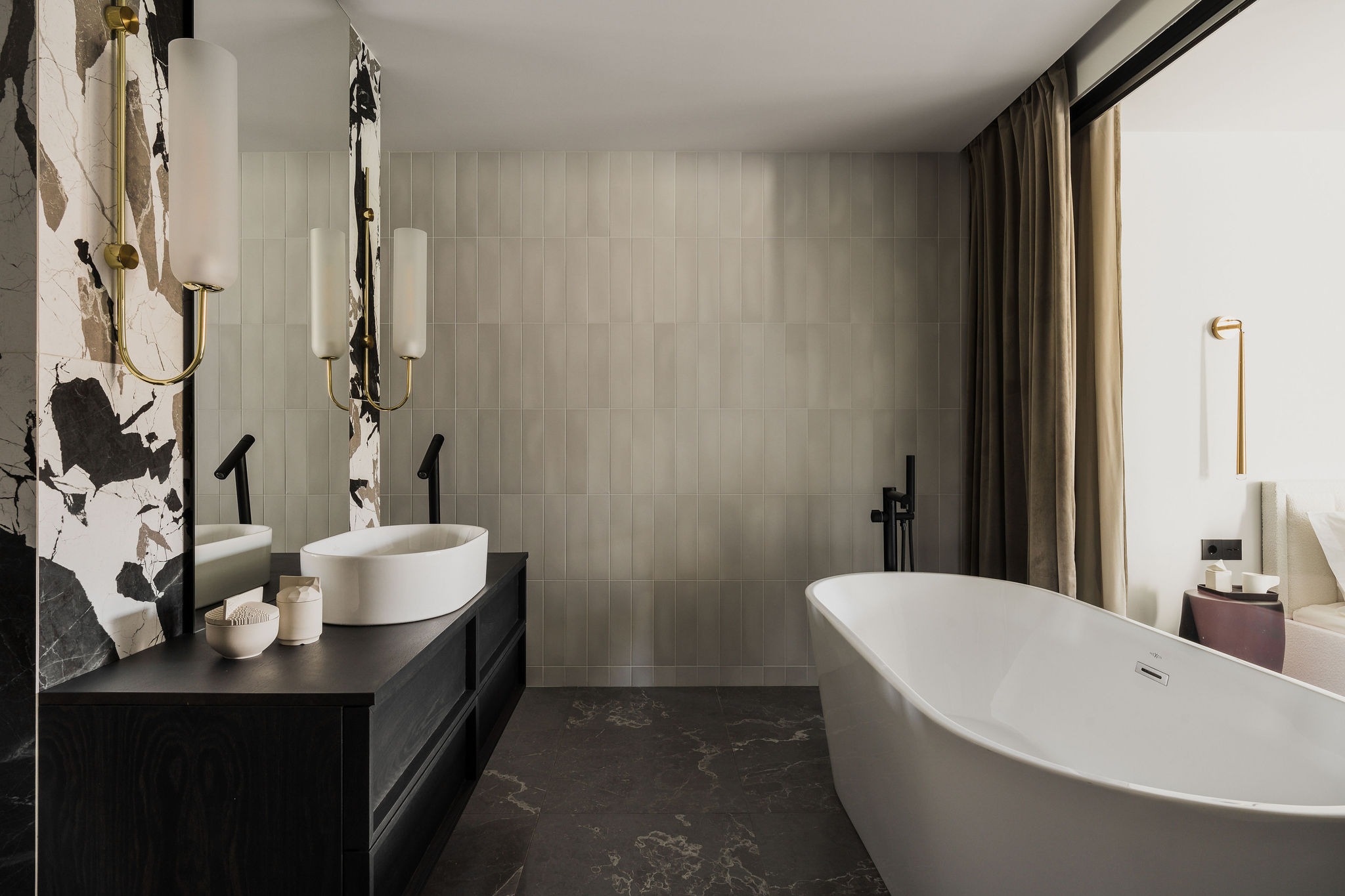 Nowoczesna łazienka z wolnostojącą wanną, umywalką nablatową na ciemnej szafce i marmurowymi akcentami, oświetlona złotymi lampami ściennymi.