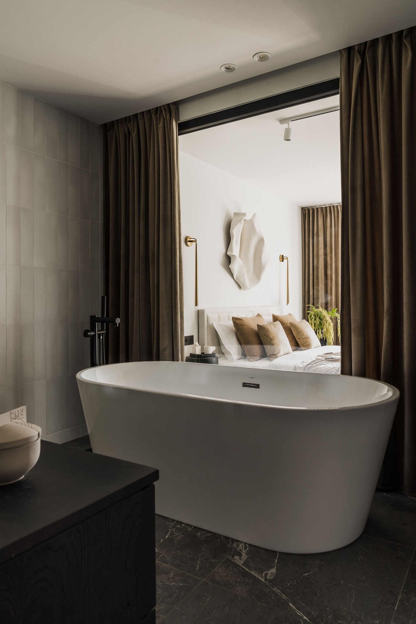 Nowoczesna wanna wolnostojąca w łazience z szarymi płytkami ściennymi i ciemną marmurową podłogą, z widokiem na sypialnię oddzieloną brązowymi zasłonami.