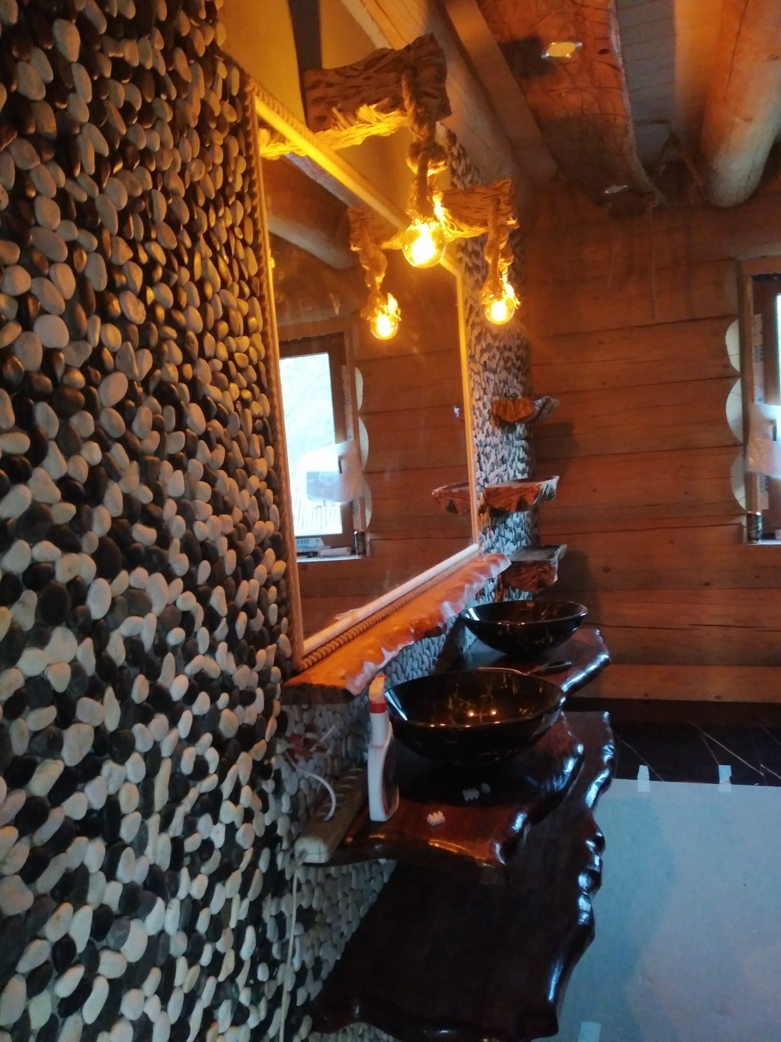 Rustykalna łazienka z drewnianymi elementami, ściana wyłożona kamieniami, czarne umywalki nablatowe i oświetlenie w stylu retro.