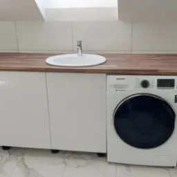 Białe szafki pod umywalką z drewnianym blatem, obok pralka Samsung, białe płytki na ścianie i marmurowa podłoga w łazience na poddaszu.