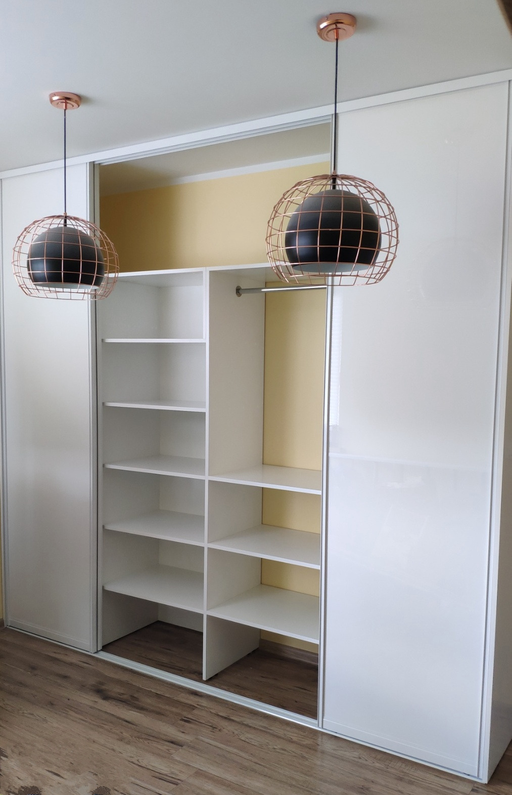 Biała szafa wnękowa z przesuwnymi drzwiami i otwartą przestrzenią z półkami, oświetlona dwoma lampami wiszącymi w stylu industrialnym z miedzianymi detalami.