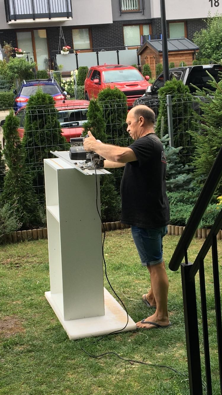 Mężczyzna w klapkach tnie płytę meblową na przenośnym stole warsztatowym na trawniku, w tle zabudowa wielorodzinna i zaparkowane samochody.